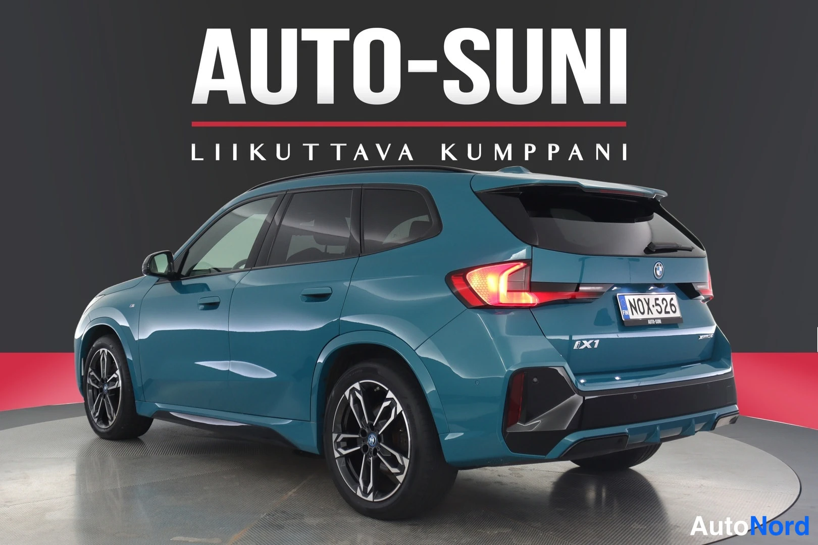 Takaviisto: Sinivihreä BMW iX1 -maasturi takaviistosta studiotaustaa vasten.