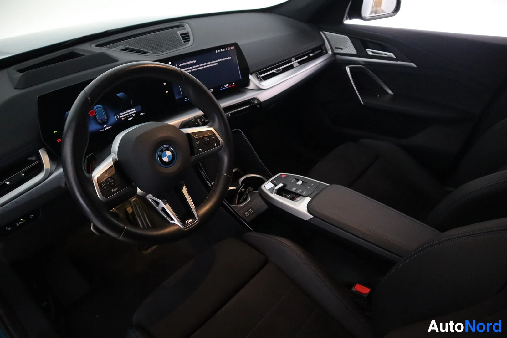 Edestä: Auton sisätilat: BMW-ohjauspyörä ja tumma kojelauta, jossa näytöt.