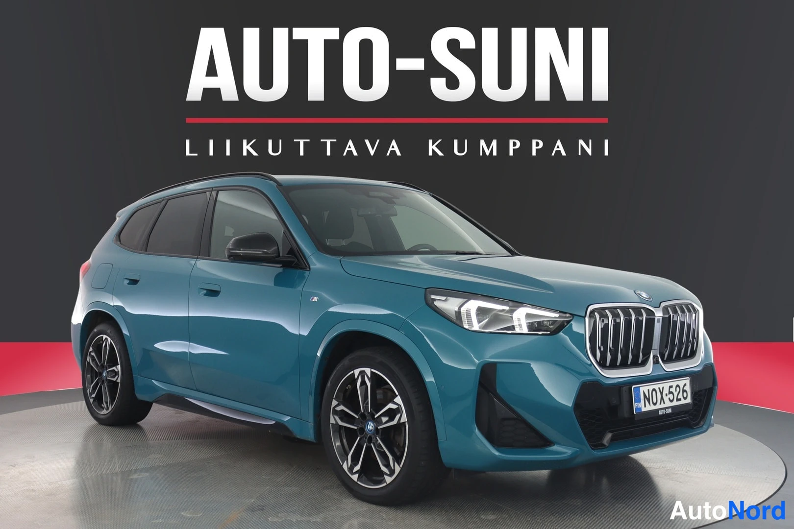 Etuviisto, nokka oikealle: Sinivihreä BMW iX1 -maasturi etuviistosta studiotaustaa vasten.