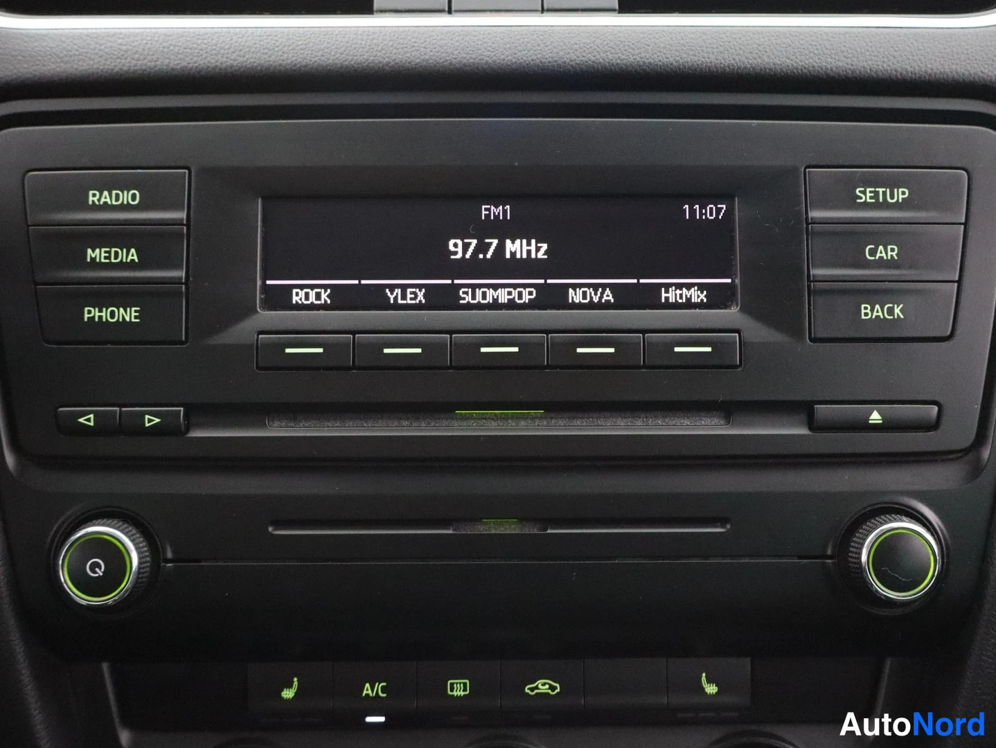 Yksityiskohta: Radio-paneeli, jossa näkyy FM1 97,7 MHz ja esiasetukset ROCK, YLEX, SUOMIPOP, NOVA, HitMix; näppäimistö RADIO, MEDIA, PHONE, SETUP, CAR, BACK.