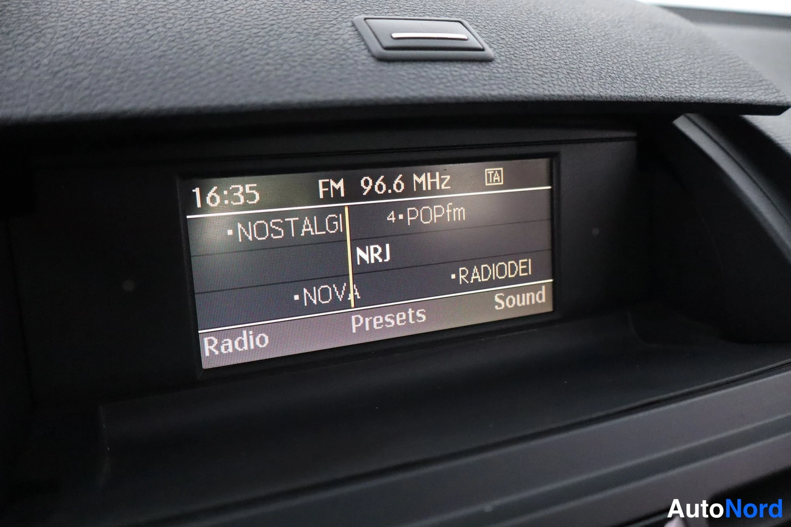 Edestä: Auton kojelaudan näyttö näyttää FM-radion 96,6 MHz kellonajalla 16:35, valikoissa Radio, Presets ja Sound.