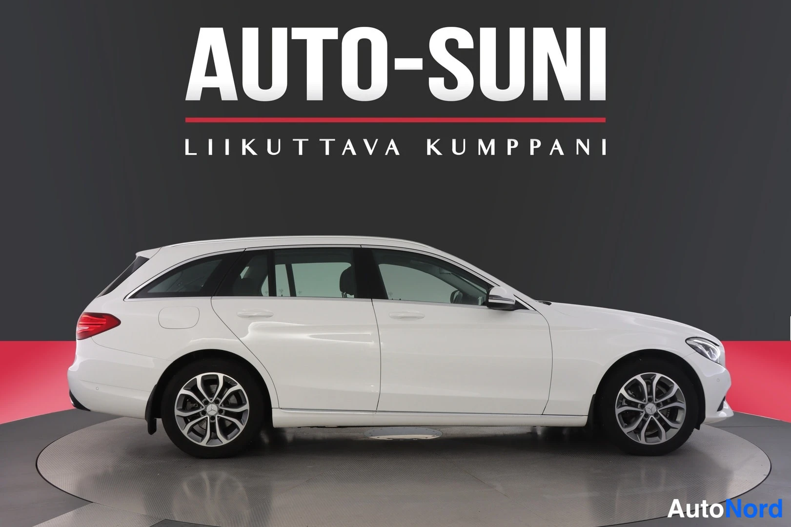 Sivulta: Valkoisen Mercedes-Benz C farmarin sivukuva vasemmalta puolelta, näyttelyalustalla.