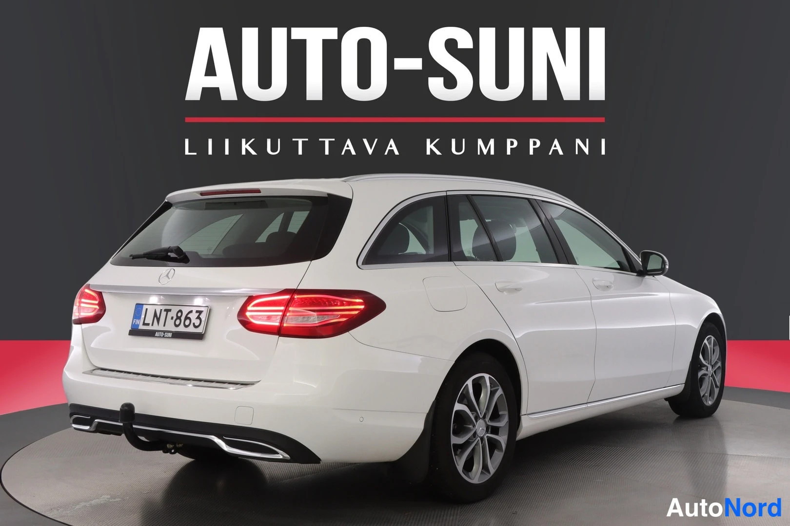 Takaviisto: Valkoisen Mercedes-Benz C farmarin takakolmisivukuva vasemmalta, punaiset takavalot, rekisteritunnus.