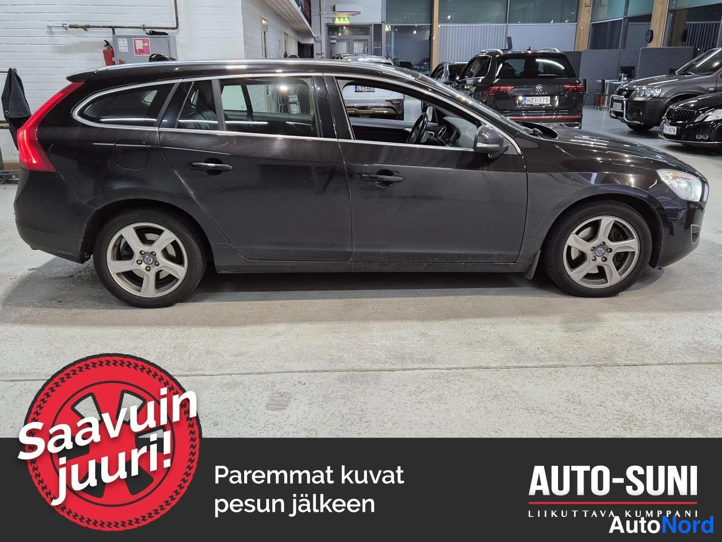 Etuviisto, nokka oikealle: Volvo V60 -farmari sivusta oikealta, tummanharmaa kori ja hopeat vanteet automyymälän sisällä.