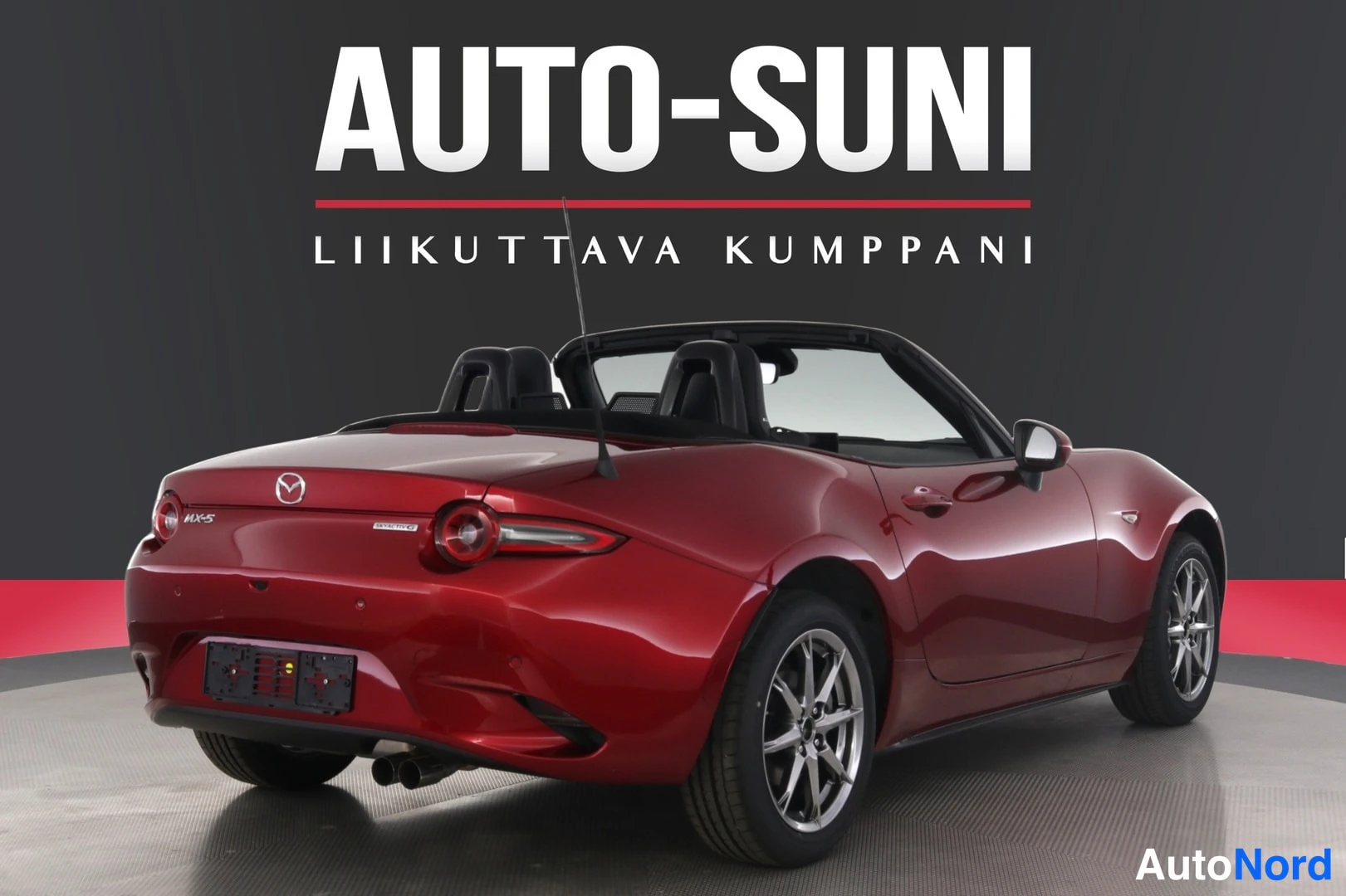 Takaviisto: Punainen Mazda MX-5 -avomalli kuvattuna takaa viistosti; kangaskatto on taitettuna, takavalot, MX-5-takamerkki ja alumiinivanteet näkyvät. Taustalla AUTO-SUNI-m
