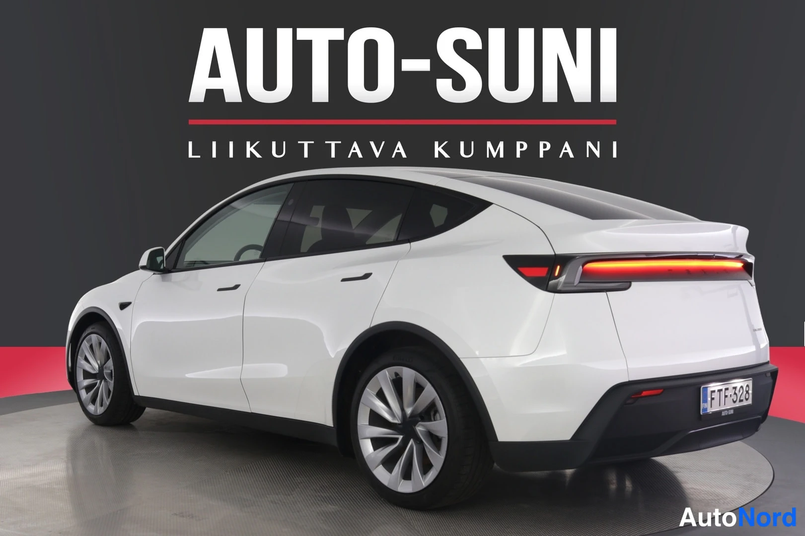 Takaviisto: Valkoinen Tesla Model Y takaa oikealta kolmesta neljäsosasta kuvattuna autosalin mustalla taustalla.