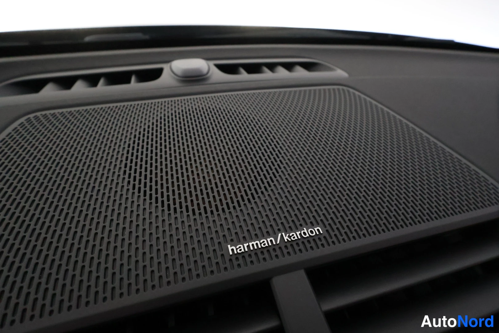 Yksityiskohta: Kojelaudan yläosan harmaa Harman Kardon -kaiuttimen ritilä, jossa merkintä 'harman/kardon' ja sen yläpuolella ilmastointilaitteen ilmaventtiiltejä.