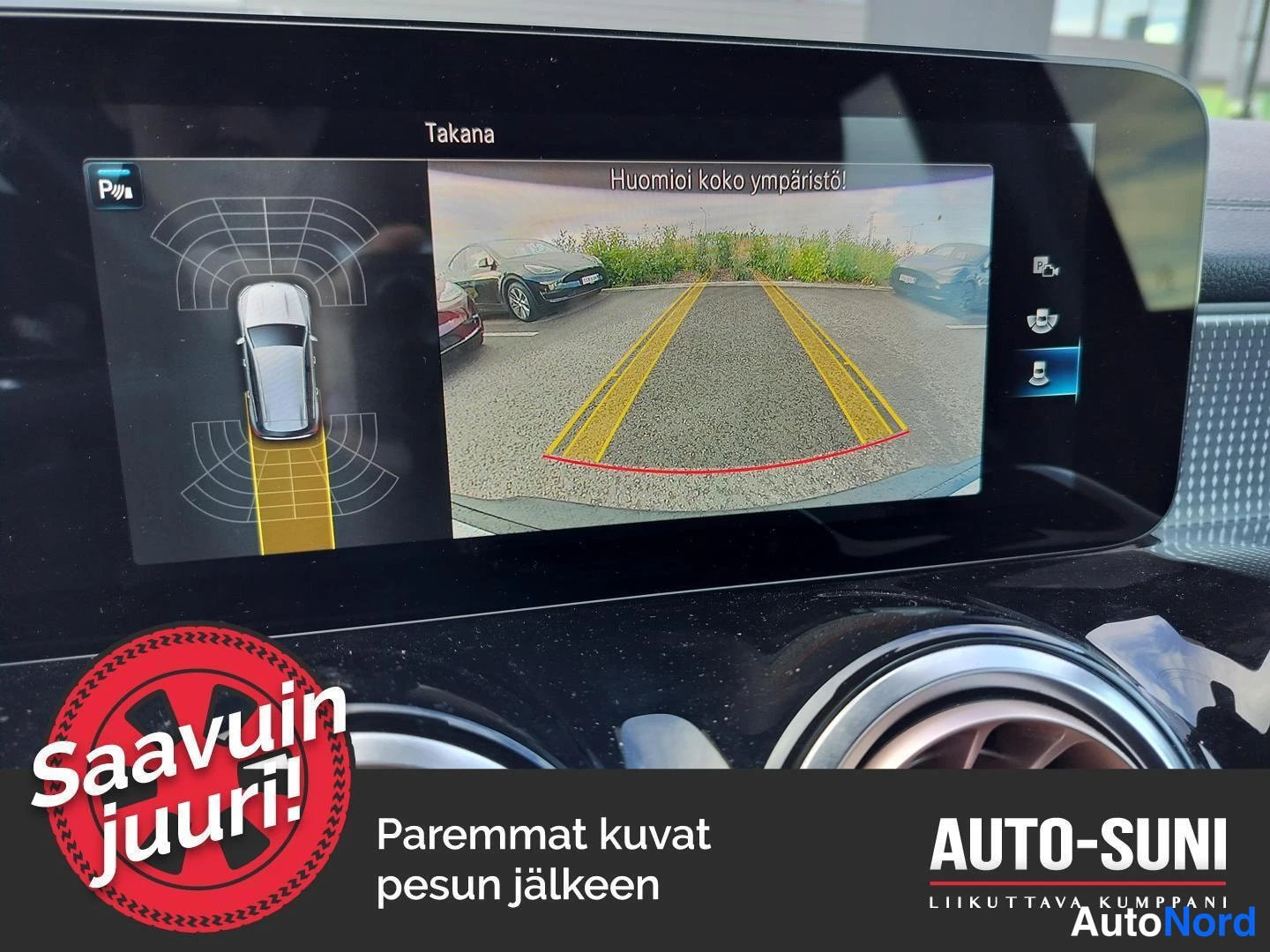 Muu: Mercedes-Benzin keskinäyttö näyttää peruutuskameran kuvan ja parkkitutkan grafiikan, tekstillä Takana ja Huomioi koko ympäristö!