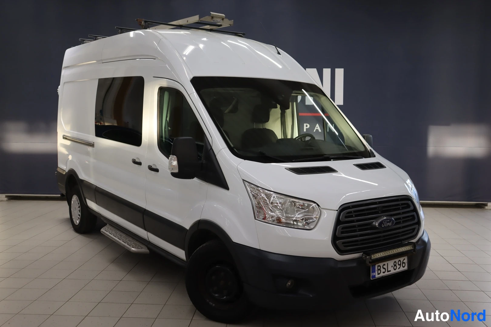 Etuviisto, nokka oikealle: Valkoinen Ford Transit -pakettiauto kuvattuna vinosti edestä oikealta; auton nokka osoittaa oikealle, kattoteline ja sivuporras näkyvät, rekisterikilpi.