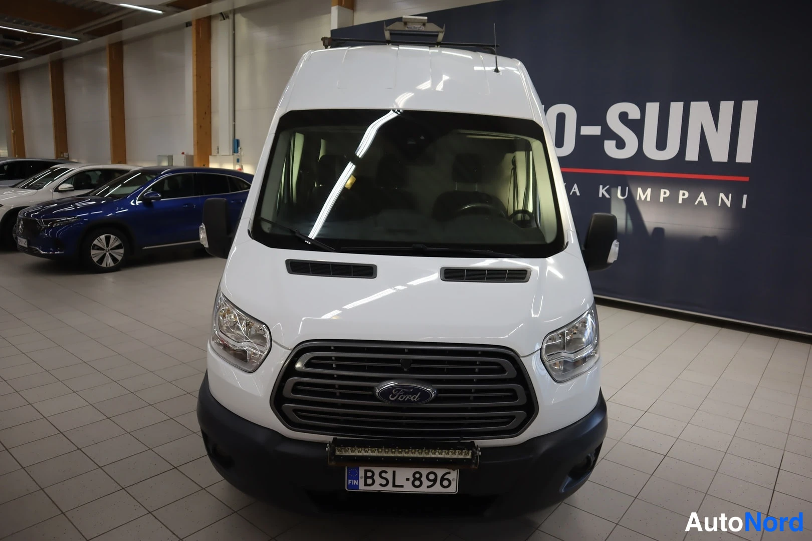 Edestä: Valkoinen Ford Transit -pakettiauto kuvattuna suoraan edestä; korkea nokka, valkoinen maalaus ja rekisterikilpi näkyvät, taustalla autotallin seinä ja mainostek
