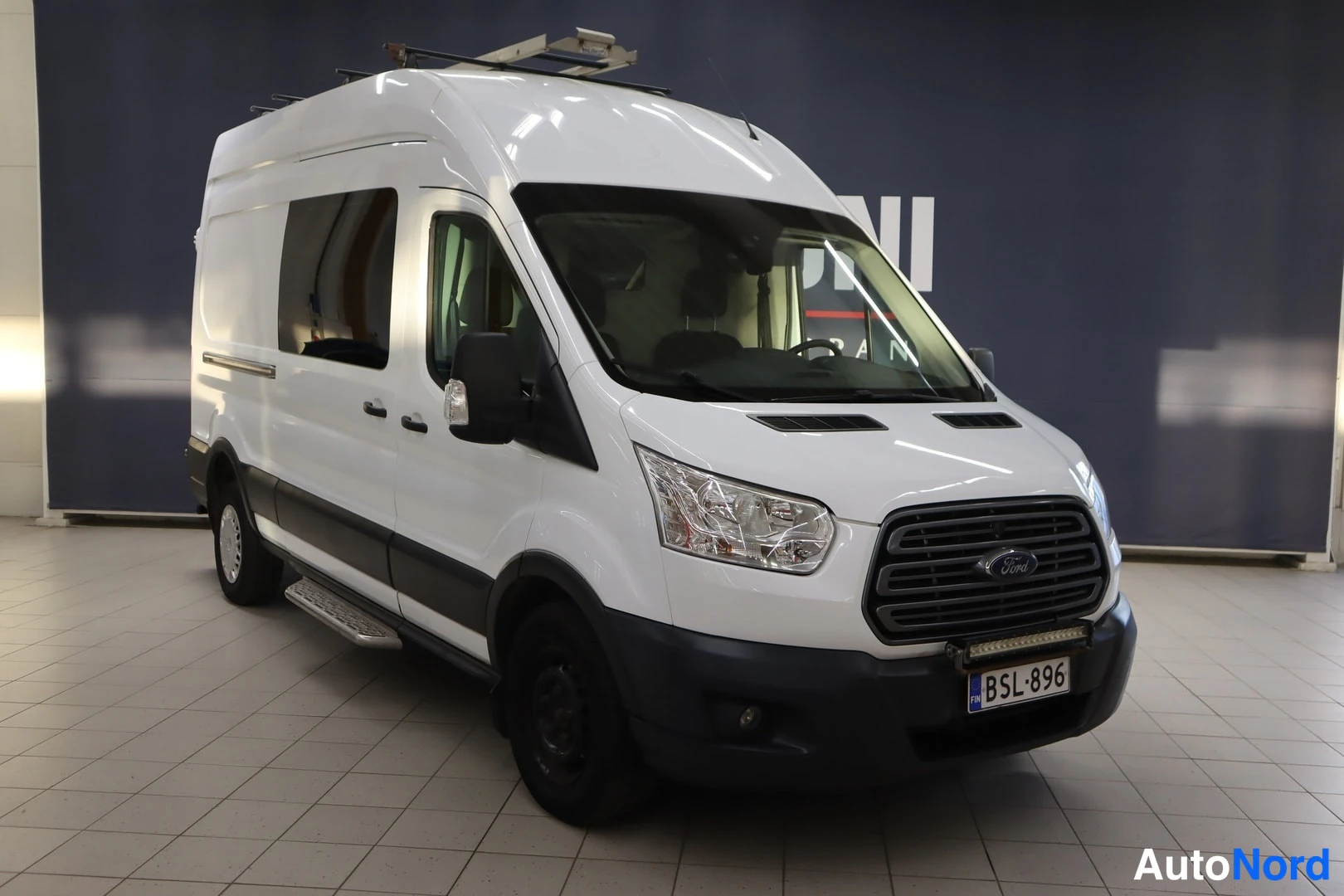 Etuviisto, nokka oikealle: Valkoinen Ford Transit -pakettiauto kuvattuna vinosti edestä oikealta autonäyttelyssä; nokka osoittaa oikealle, kattoteline, sivuporras ja rekisterikilpi