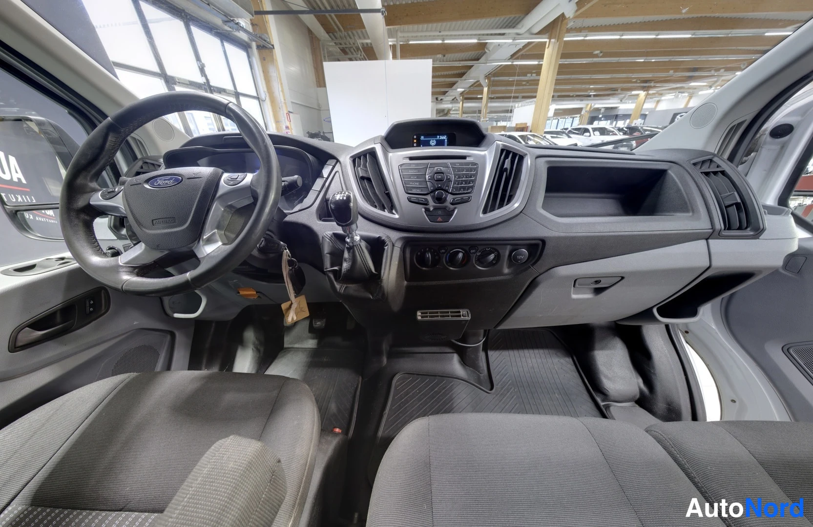 Muu: Ford Transitin sisätilat: ohjaamo, manuaalivaihteen vaihdekeppi, radio ja tummat kankaistuolit.