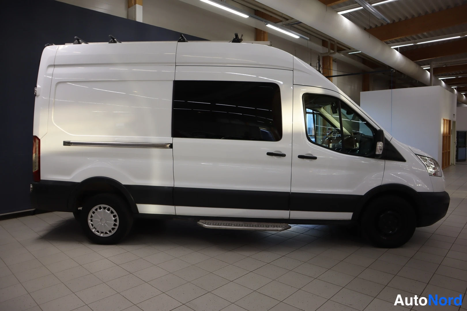 Etuviisto, nokka oikealle: Ford Transit -pakettiauton etukulma oikealta, valkoinen koripohja, sivuporrashenge ja kattoteline näkyvät.