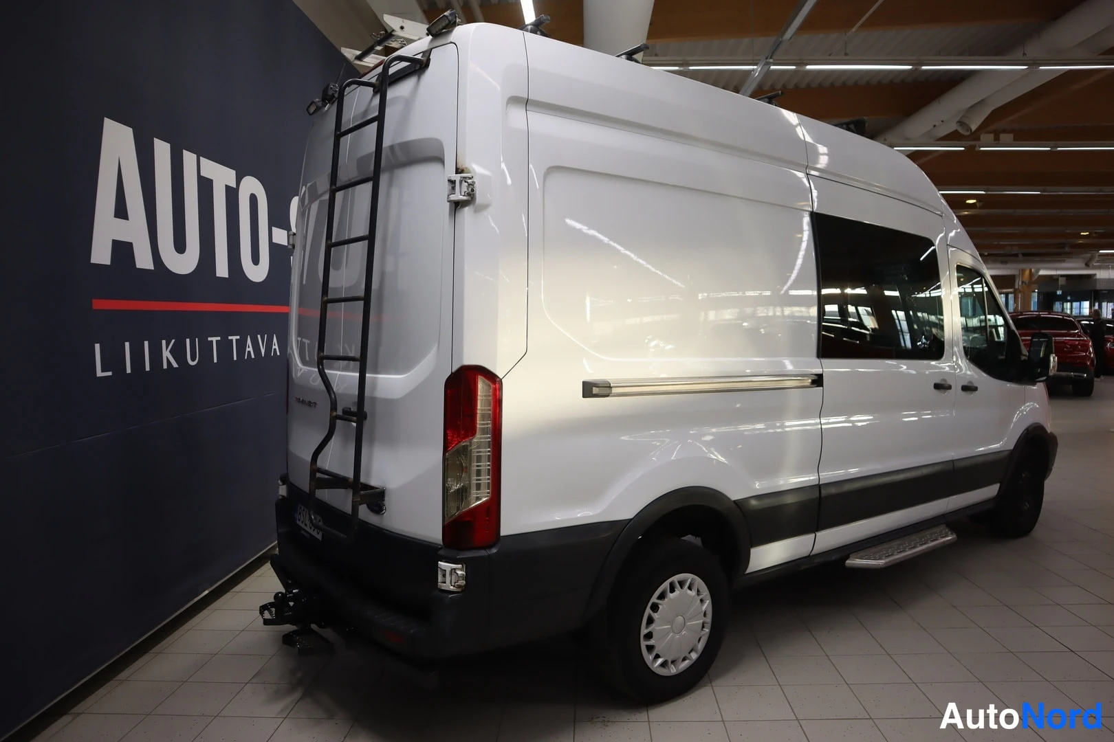 Takaviisto: Ford Transit -pakettiauton takakulma vasemmalta, peräladder, kattoteline ja vetokoukku näkyvät.