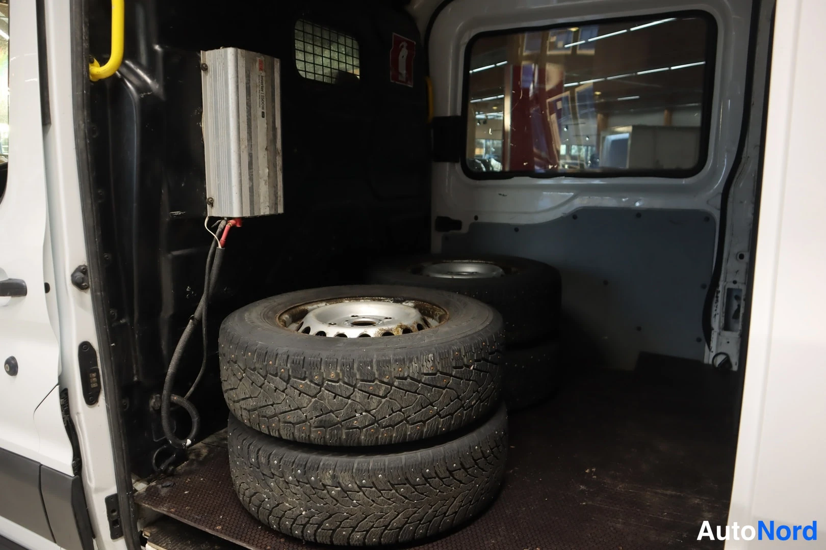 Sivulta: Ford Transitin tavaratilan sisäkuva, lattialla neljä nastarenkaita vanteilla ja seinällä kiinnitetty laite.