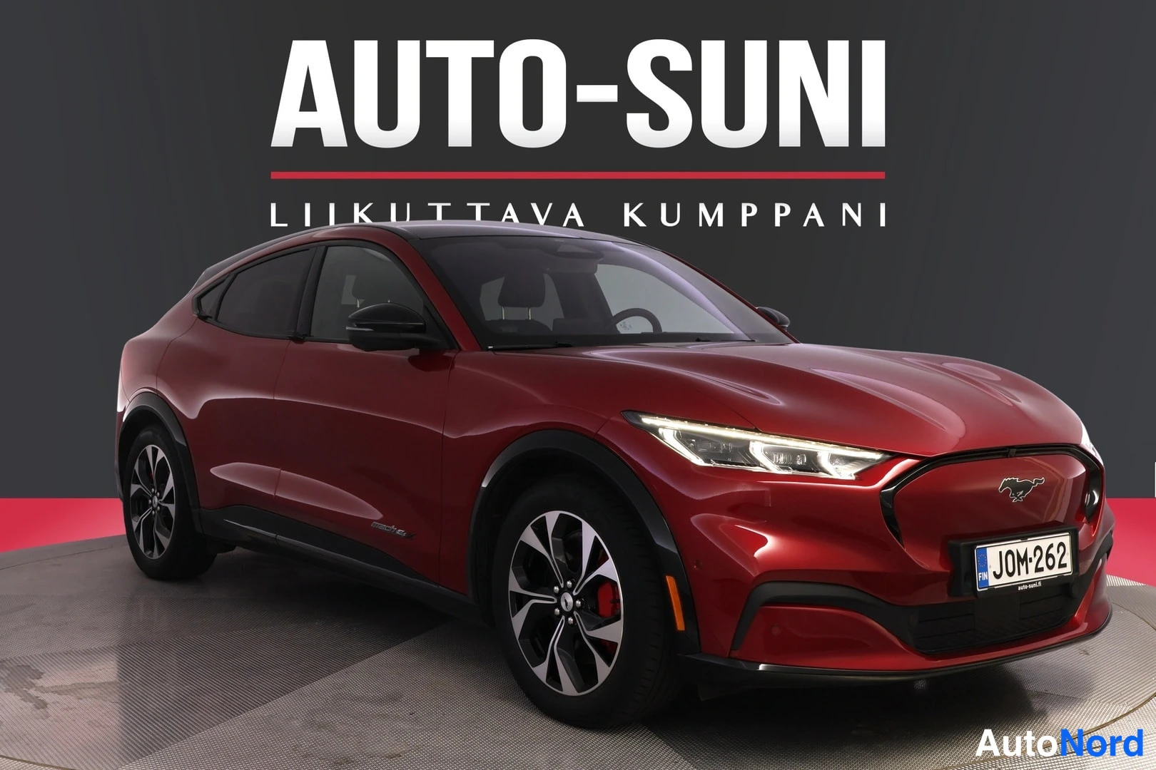 Etuviisto, nokka oikealle: Punainen Ford Mustang Mach-E -maasturikupé kuvattuna kolmesta neljäsosan etukulmasta, rekisterikilpi, studiothanen tausta ja AUTO-SUNI-logo.