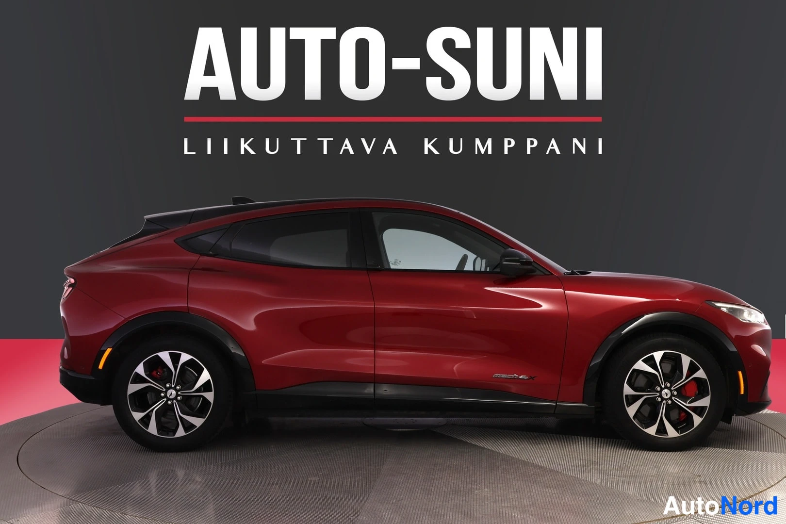 Sivulta: Sivuprofiili punaisesta Ford Mustang Mach-E -autosta, musta katto ja kevytmetallivanteet, taustalla tumma seinä ja AUTO-SUNI-teksti.
