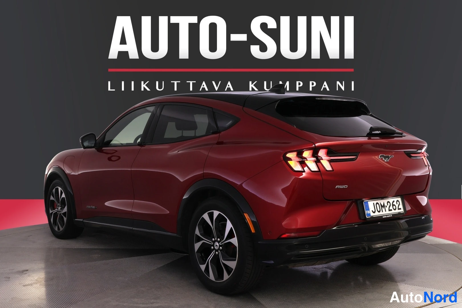 Takaviisto: Punainen Ford Mustang Mach-E takaa kolmesta neljäsosan kulmasta, musta katto ja takavalot päällä, rekisterikilpi, AUTO-SUNI-tausta.