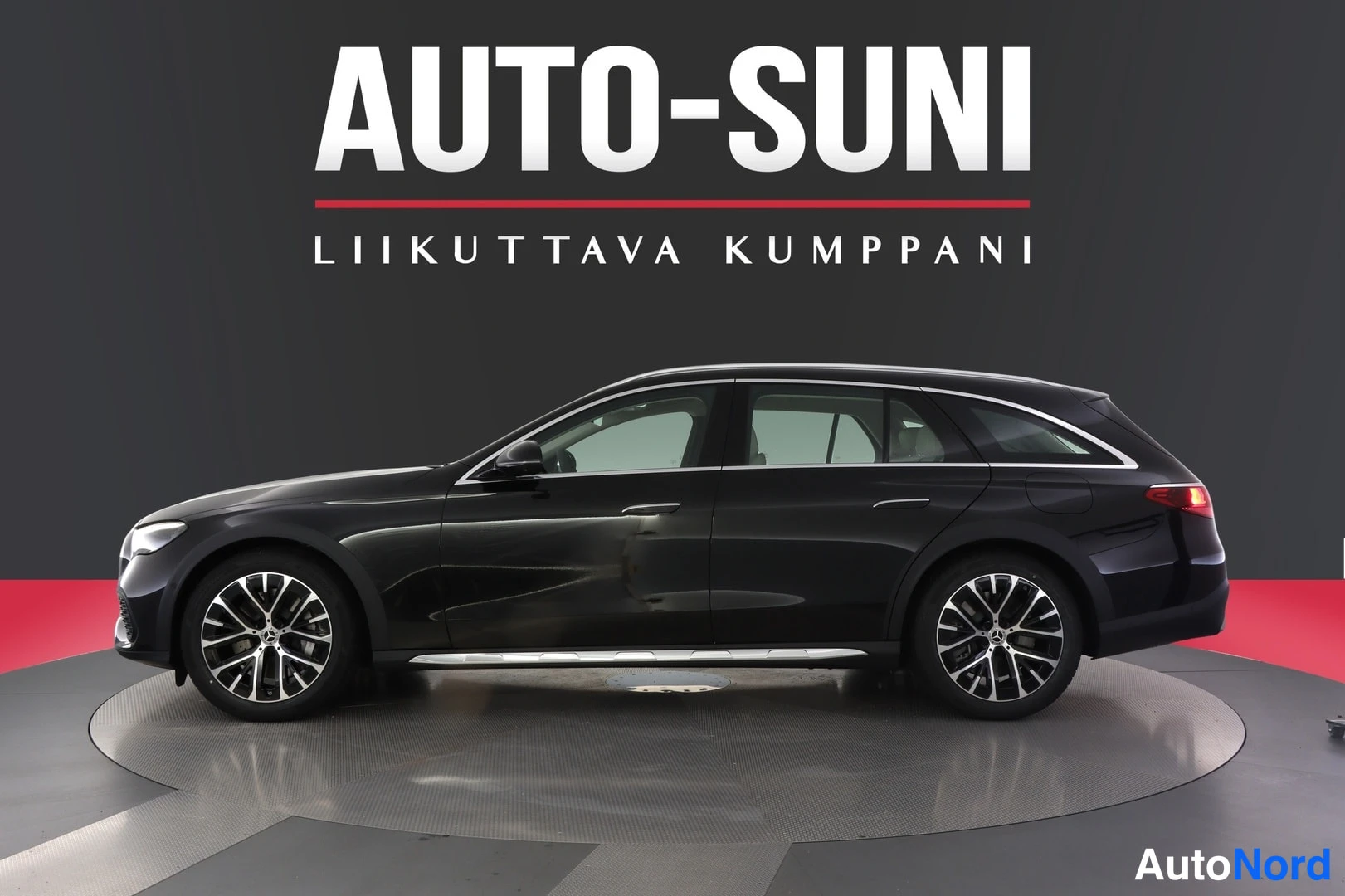 Takaviisto: Musta Mercedes-Benz farmariauto kuvattu takaa sivulta myyntihallin pyörälle asetettuna, taustalla AUTO-SUNI-kyltti.