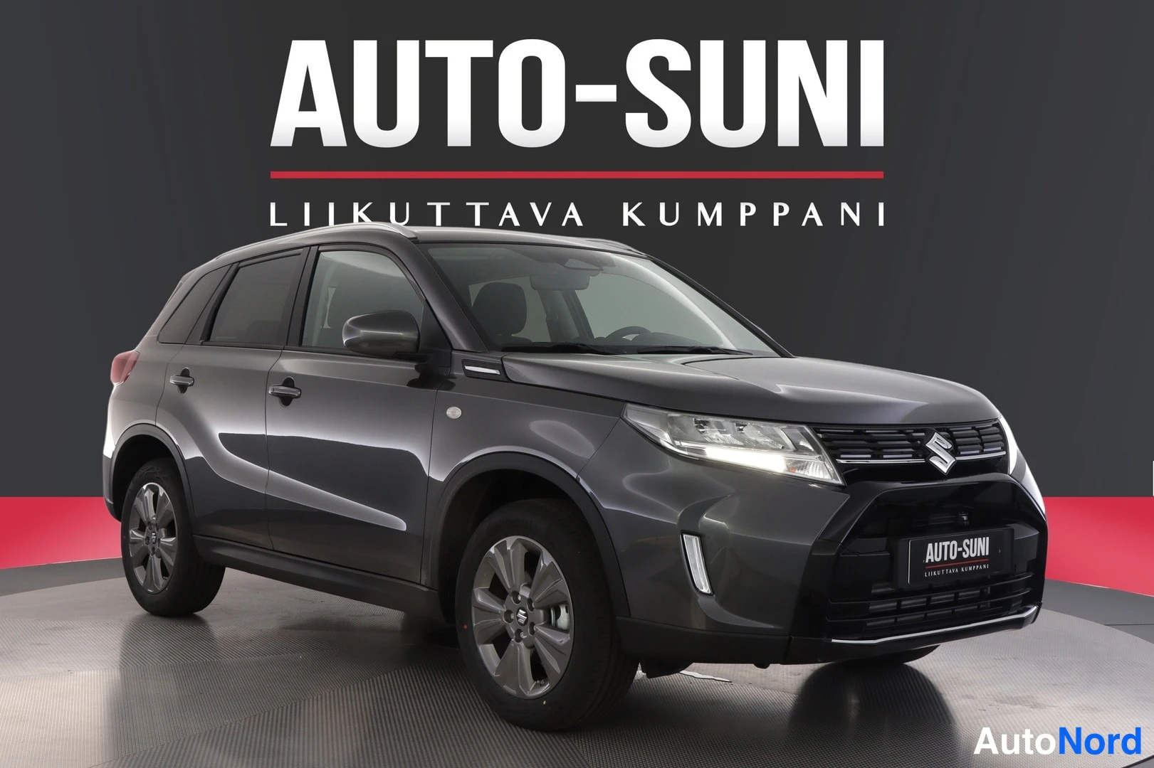 Etuviisto, nokka oikealle: Tummanharmaa Suzuki Vitara studio-aiheisessa valokuvassa, edestä vasemmalta kolmasosa-kulmasta. Auton etupää, etusuoja ja kylkipaneelet näkyvät, valkoi