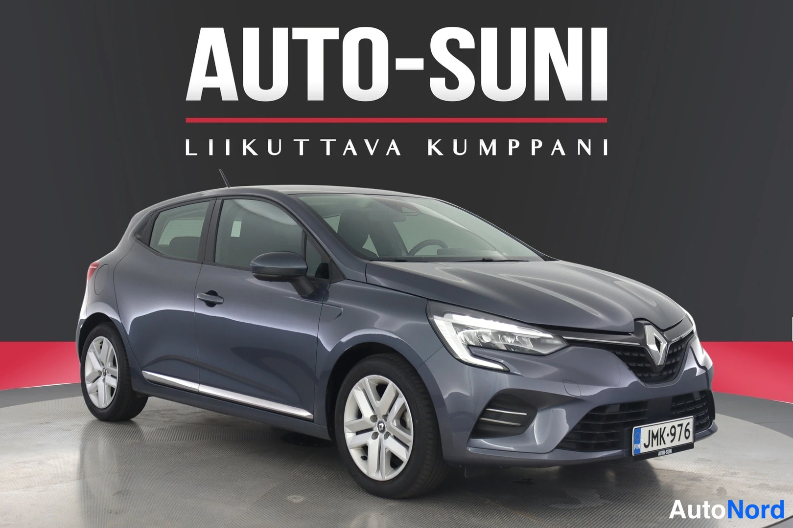 Etuviisto: Sinertävän tummanharmaa Renault Clio -viistoperä esillä myyjän taustakyltin edessä, kolmen neljäsosan etunäkymä vasemmalta; LED-ajovalot, sumuvalot ja kevytmeta