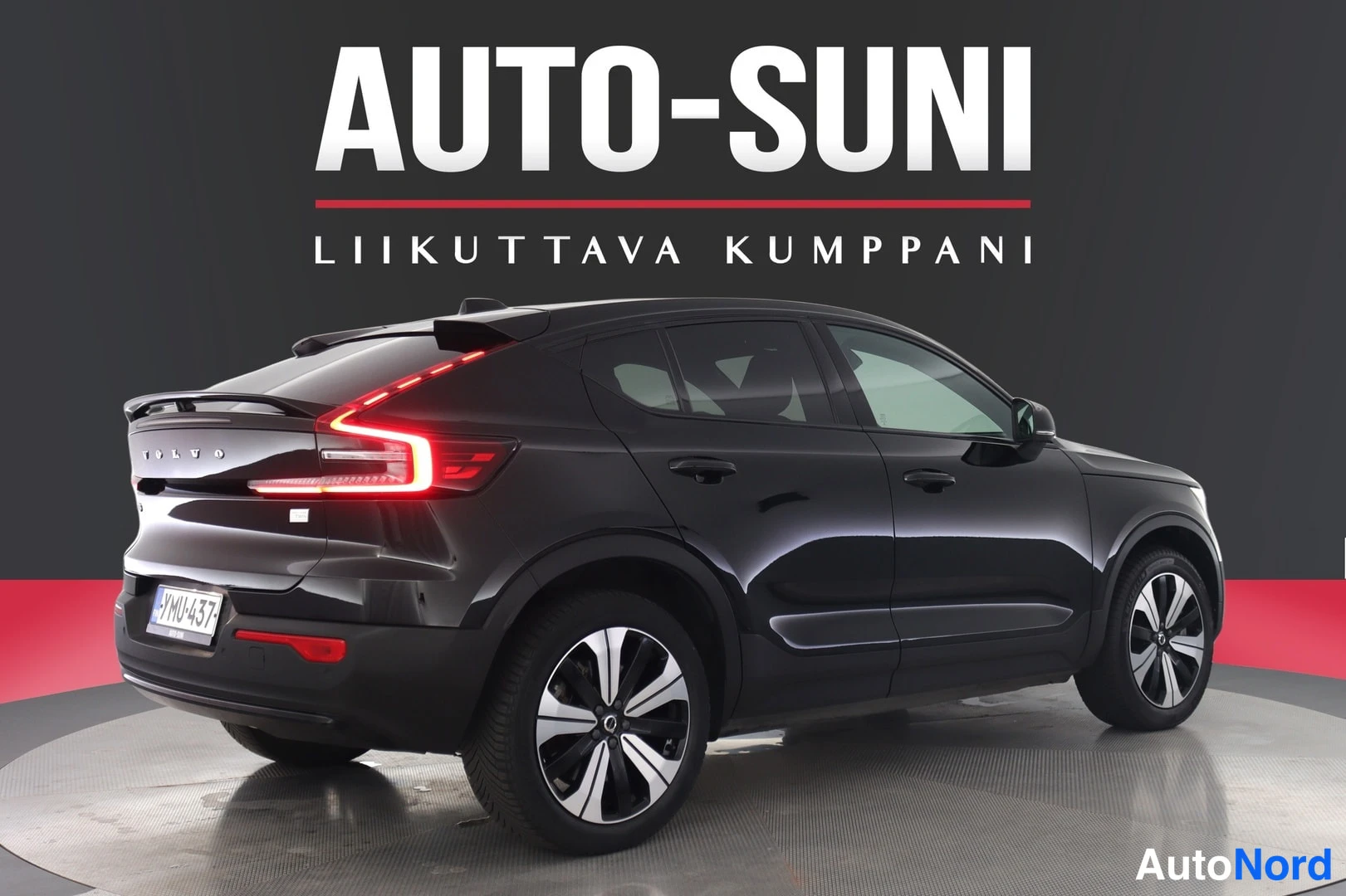 Takaviisto: Mustan henkilöauton takaosa ja oikea kylki valaistuilla takavaloilla, rekisteri.