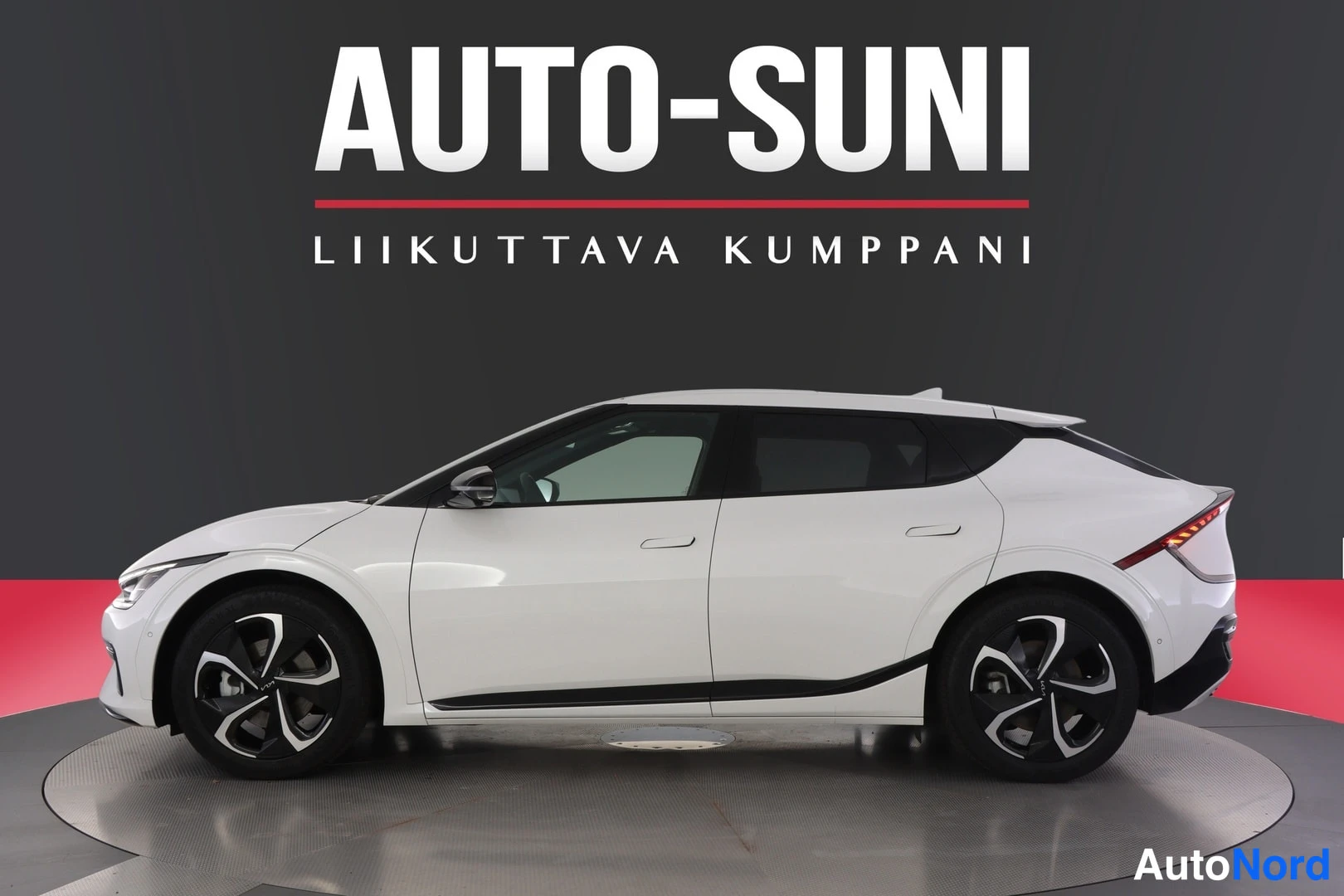 Sivulta: Ajomoneuvon profiilinäkymä studio: valkoinen crossover-tyylinen ajomoneuvo, keula vasemmalla, mustat pyörät ja tummat ikkunat, tausta harmaa-punainen ja teksti