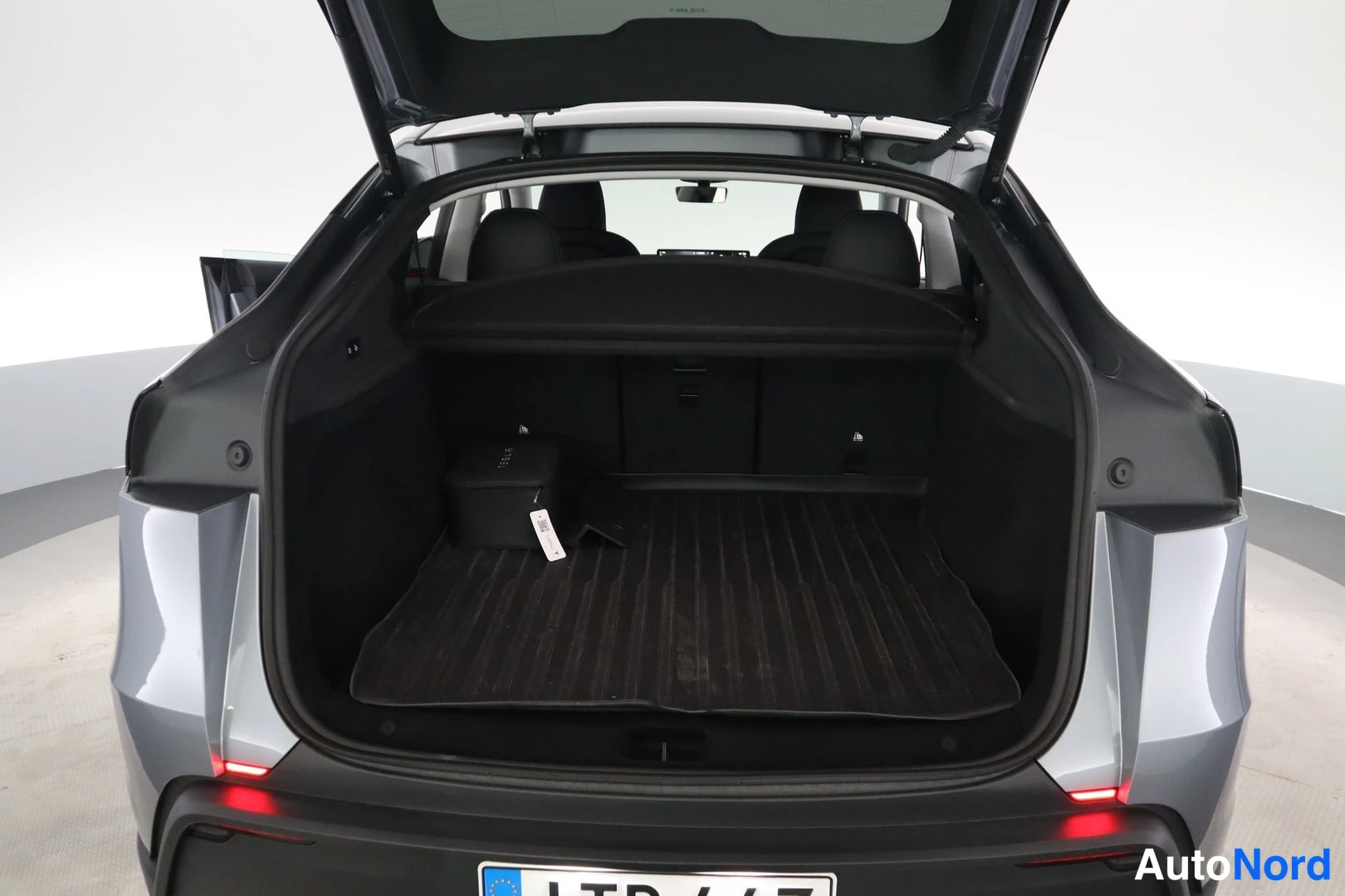 Takaa: Tesla Model Y takaa kuvattuna, tavaratilan luukku auki: tumma tavaratilan matto ja musta säilytyslaatikko tavaratilassa.