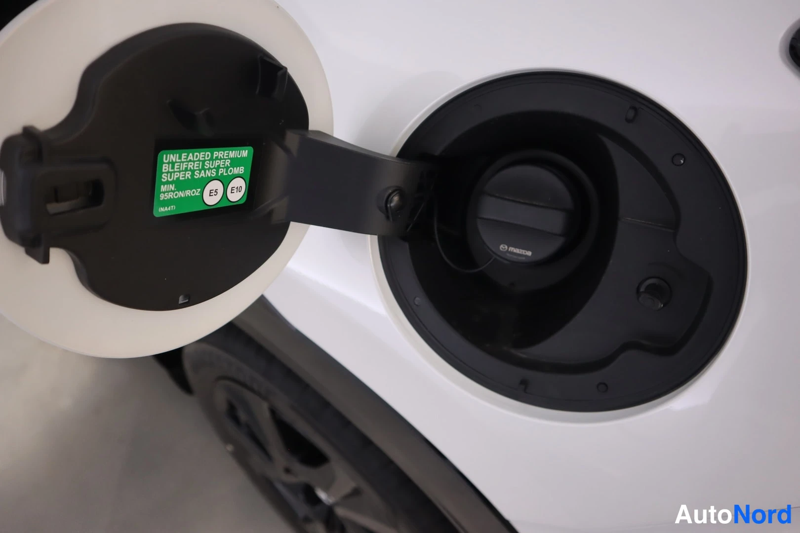 Yksityiskohta: Avattu polttoainetäyttöaukko valkoisen auton sivussa; mustassa luukussa vihreä tarra tekstillä UNLEADED PREMIUM, BLEIFREI SUPER, SUPER SANS PLOMB, MIN. 95RON/RO