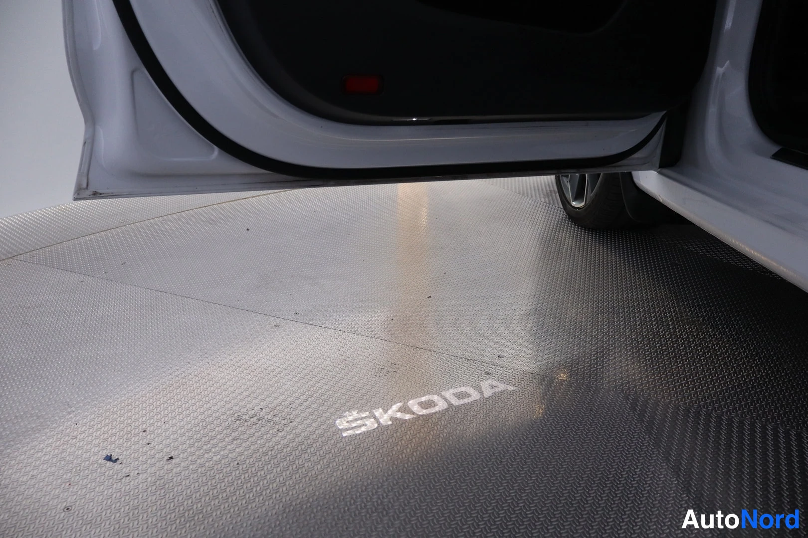 Edestä: Oviaukon alapuoli Skoda-merkinnällä lattiassa; valkoinen auton ovipellin ja takapyörän osa näkyvät.