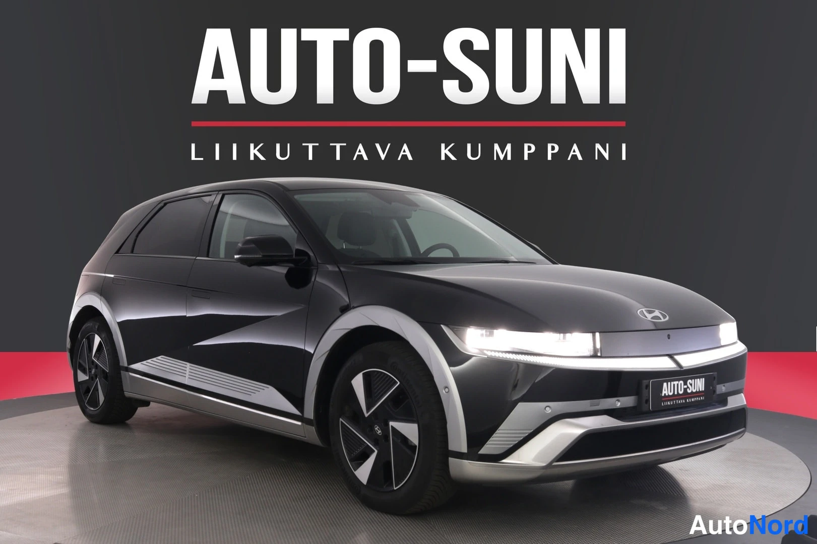 Etuviisto, nokka oikealle: Musta Hyundai IONIQ 5 -henkilöauto kulmassa edestä vasemmalta kuvattuna myymälä Näyttelytilassa AUTO-SUNI-logon alla.