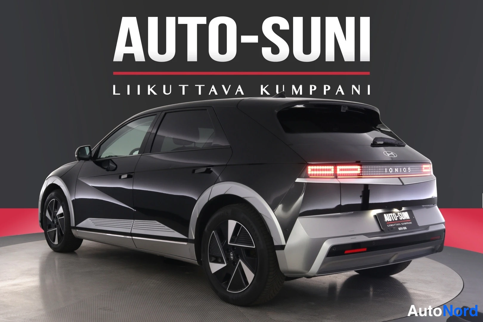 Takaviisto: Musta Hyundai IONIQ 5 -henkilöauto takaa oikealta kuvattuna Näyttelytilassa AUTO-SUNI-logon alla.