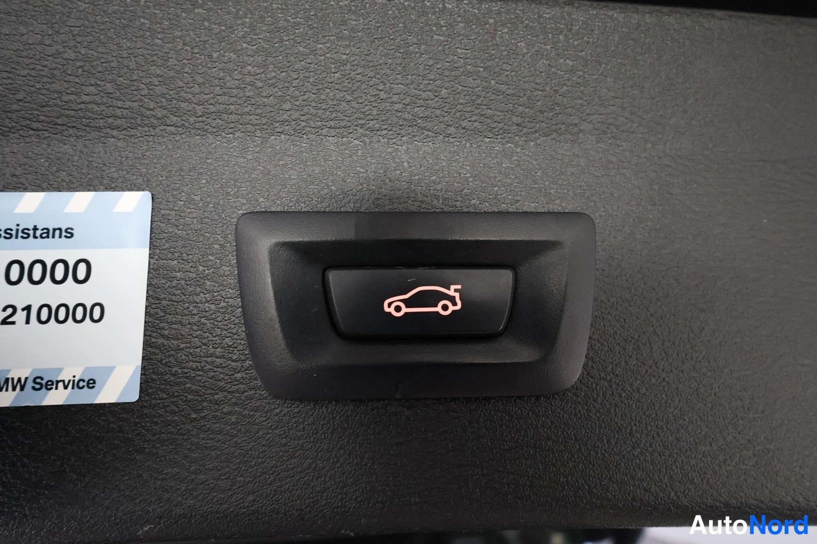 Yksityiskohta: Close-up of the fuel filler door release button located on the left side panel below the dashboard. The button displays a red icon of a car with an open fuel ca