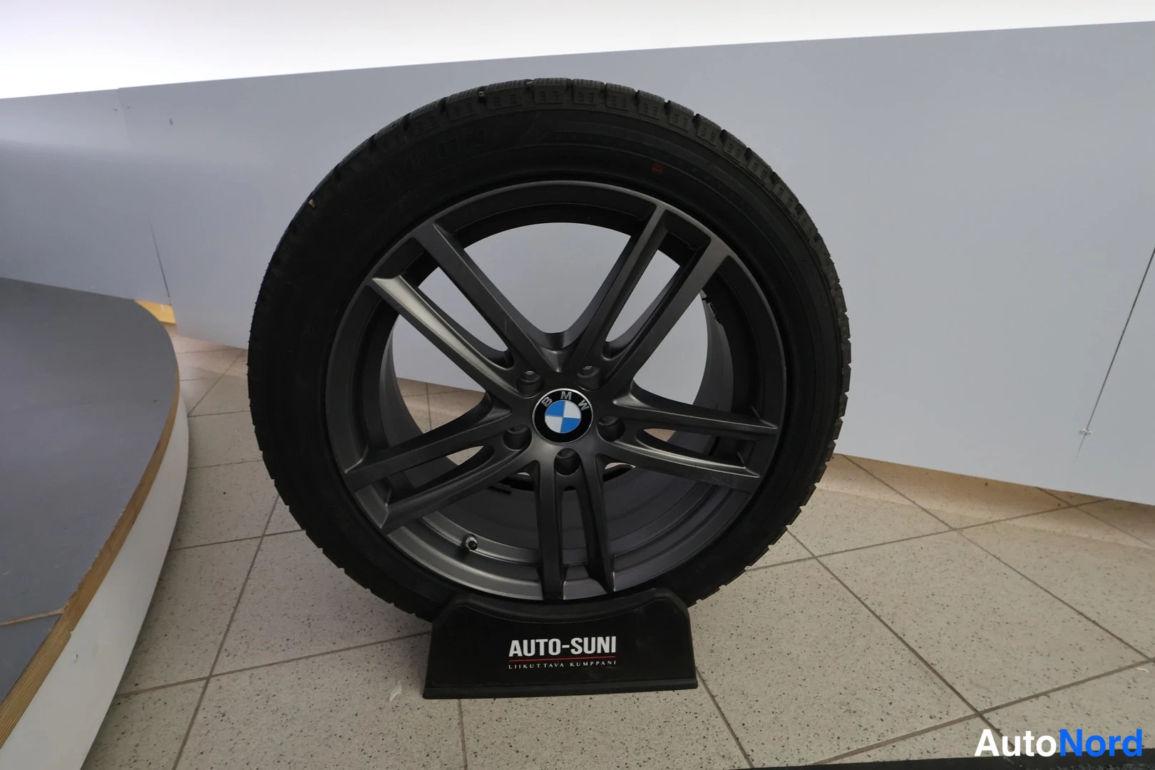 Yksityiskohta: BMW-merkkinen tumma kevytmetallivanne renkaalla AUTO-SUNI-näytetelineen päällä sisätilassa.