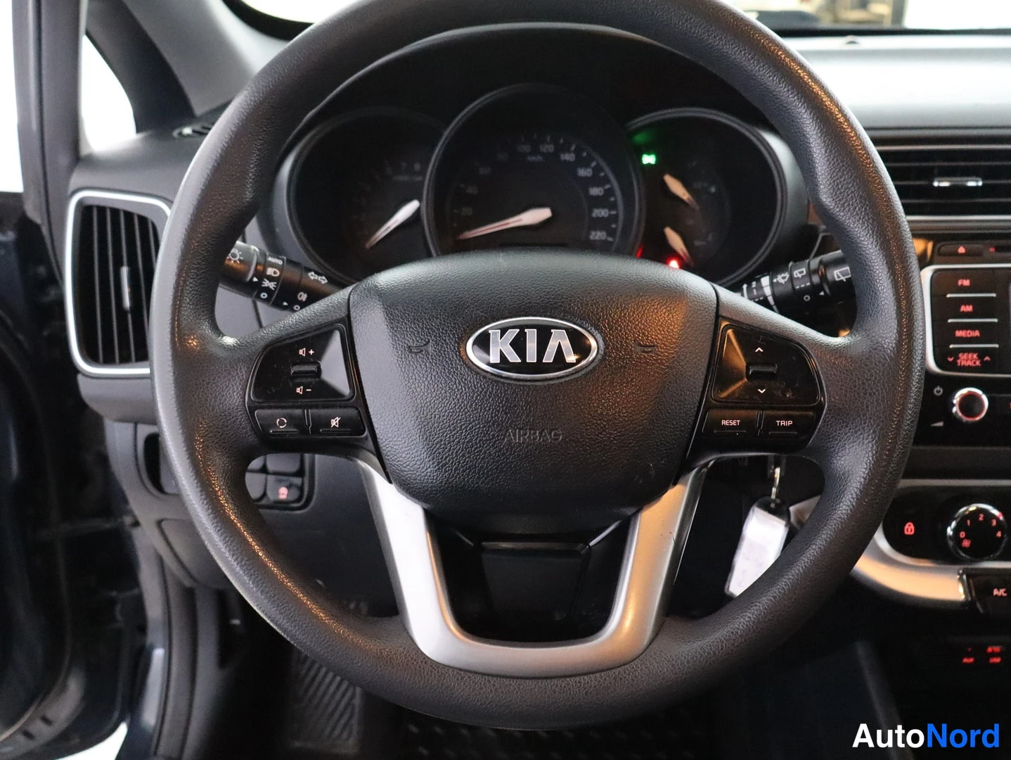 Muu: Kia Rio -henkilöauton ohjauspyörä KIA-logolla ja AIRBAG-tekstillä, taustalla kojetaulu ja keskikonsolin radiopaneeli.