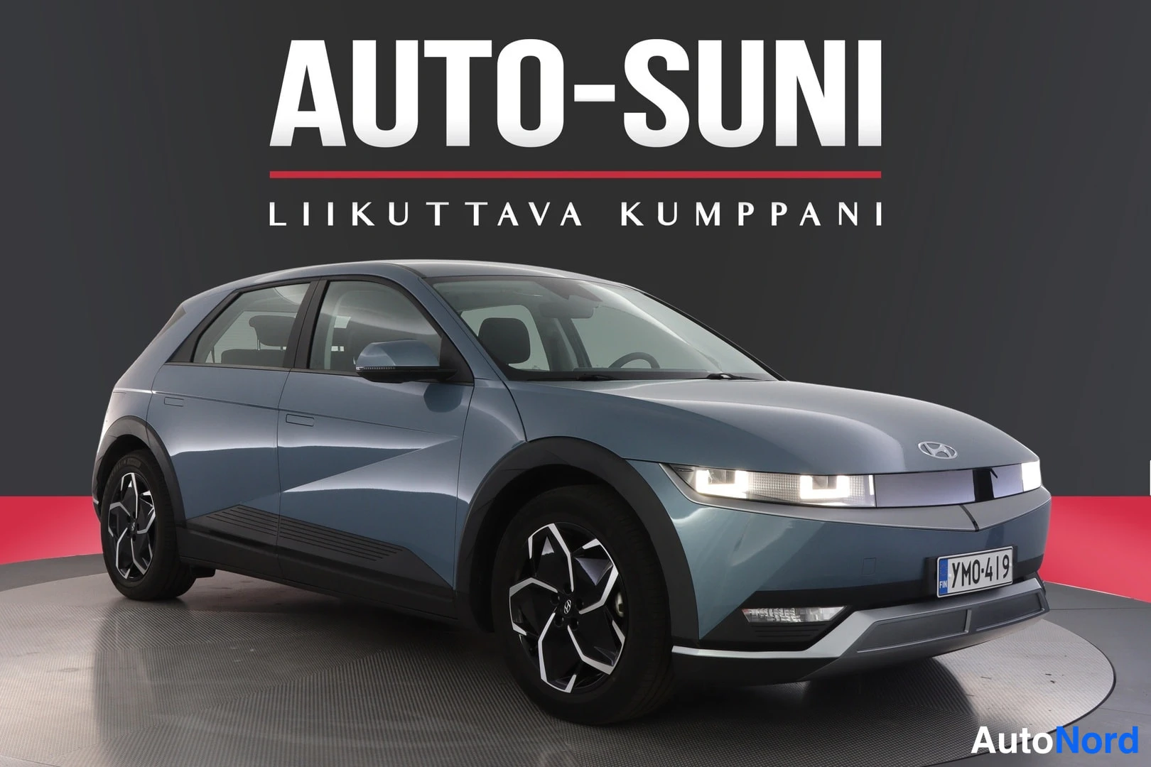Etuviisto, nokka oikealle: Blue Hyundai IONIQ 5 parked on a rotating platform in a studio. Front and driver's side view with headlights illuminated.