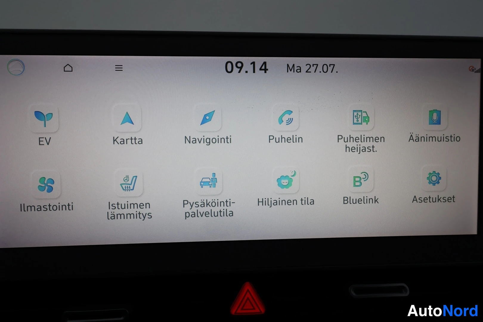 Yksityiskohta: Car infotainment touchscreen displaying home menu with apps for EV, navigation, phone, climate control (Ilmastointi), heated seats (Istuimen lämmitys), parking,
