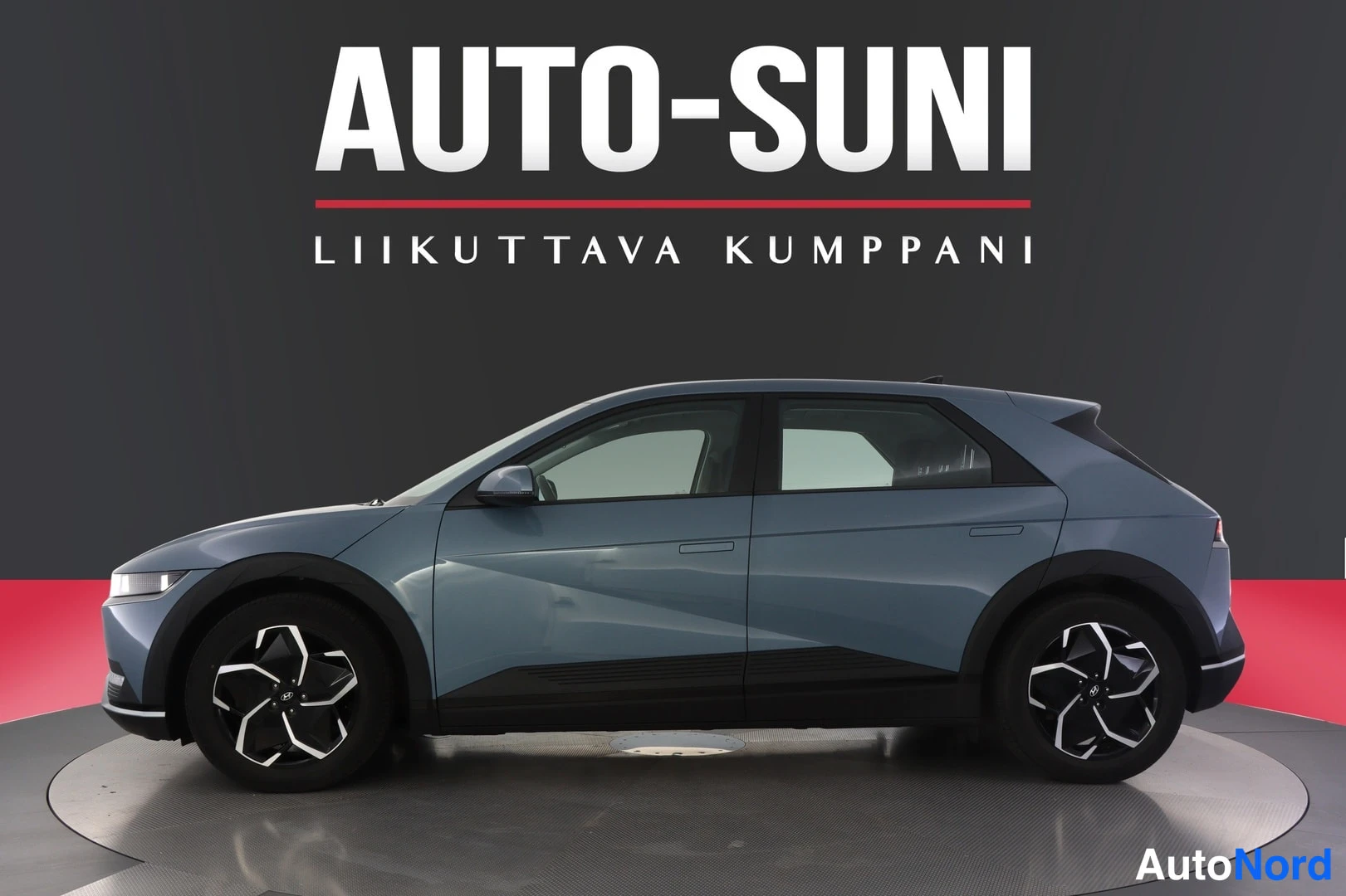 Sivulta: Hyundai IONIQ 5:n sivuprofiili vasemmalta; korin väri on harmaansininen, taustalla harmaa seinä ja AUTO-SUNI-teksti.