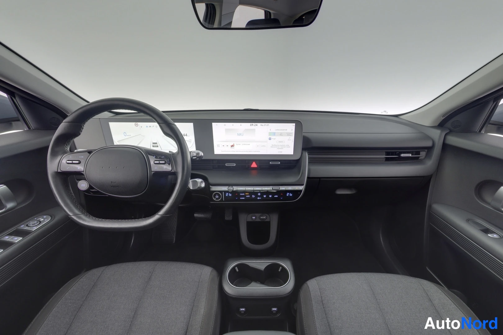 Edestä: Hyundai IONIQ 5:n sisätilat kuvattuna takaosalta kohti kojelautaa: ohjauspyörä vasemmalla, digitaalinen mittaristo ja suuri keskikonsolin kosketusnäyttö karttan
