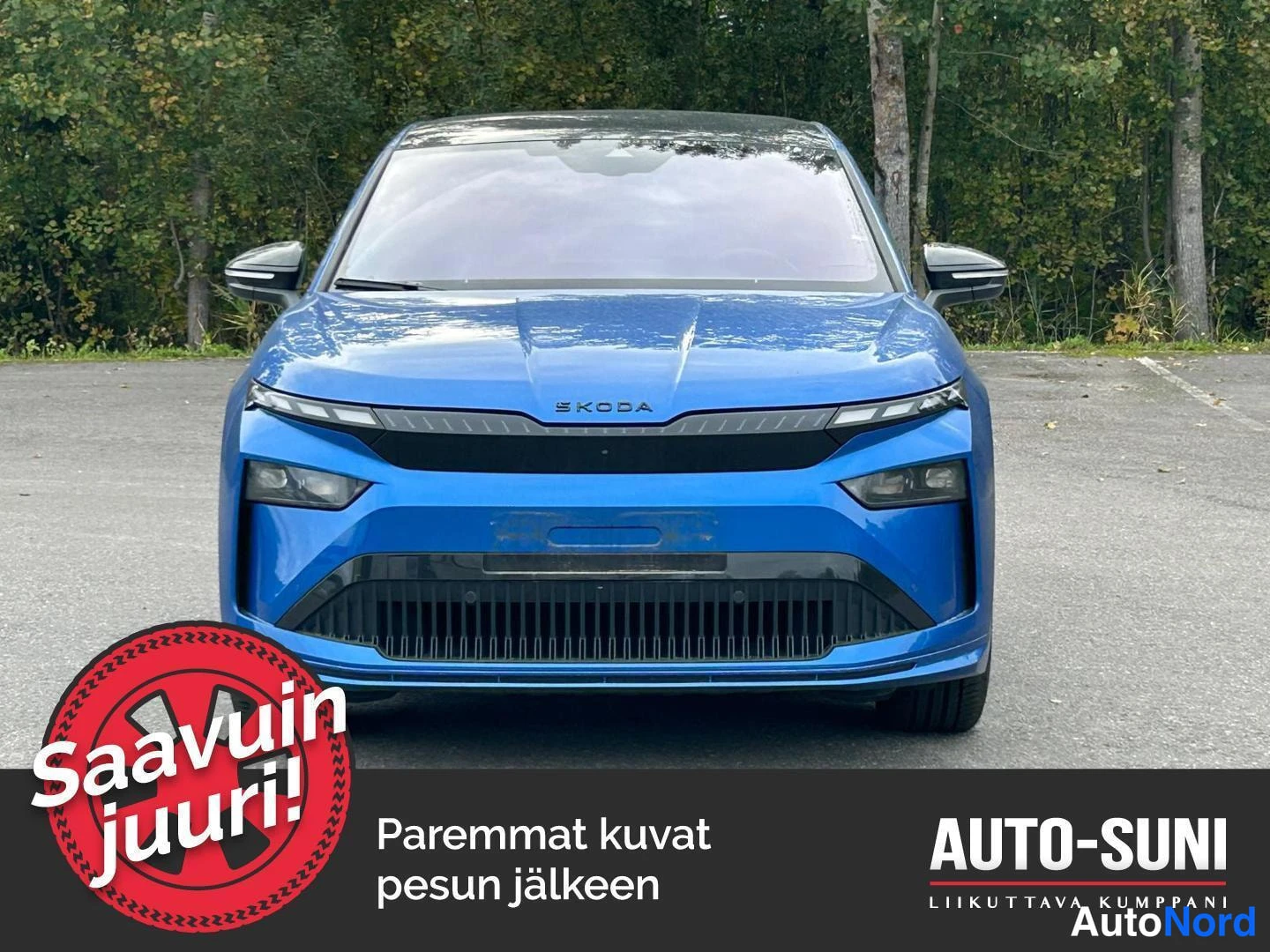 Edestä: Ensinäkökuva kirkansinisestä Škoda Enyaqista etusäleikkönä ja valaisimilla puuston edessä.