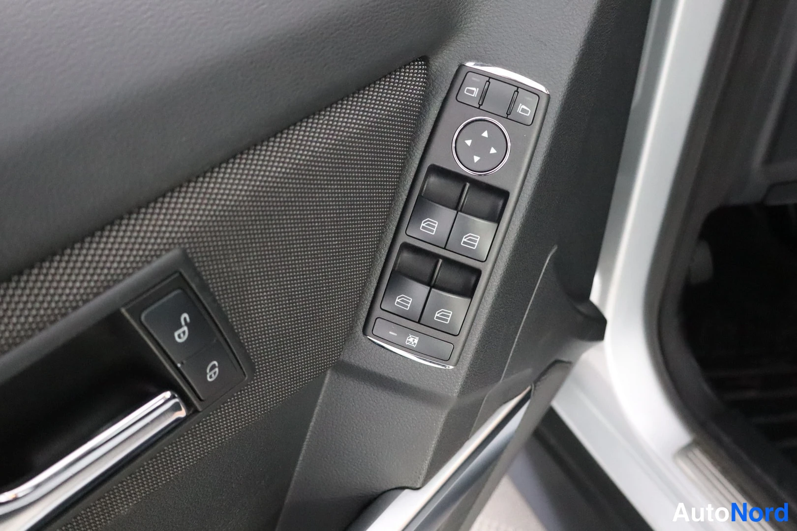 Yksityiskohta: Close-up of the driver's side door panel controls. Visible elements include buttons for window operation (up/down/left/right), central locking (lock/unlock symb