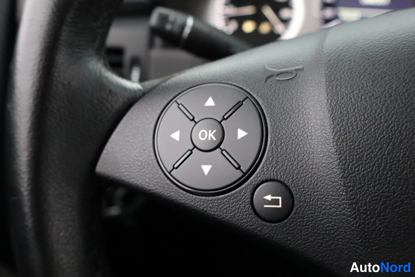Yksityiskohta: Close-up of the left-hand spoke of a black steering wheel featuring directional arrow buttons around an 'OK' button and a return arrow icon.