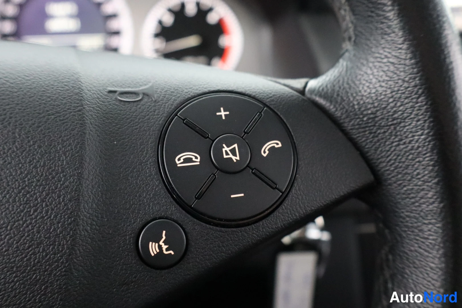 Yksityiskohta: Close-up of the right-hand spoke of a black steering wheel with audio and phone control buttons including volume adjustment symbols and handset icons.