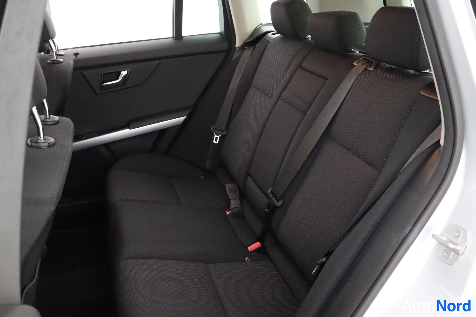 Takaistuimet: Rear passenger seating area viewed from an open rear door showing black cloth upholstery, three headrests and a metal grab handle.