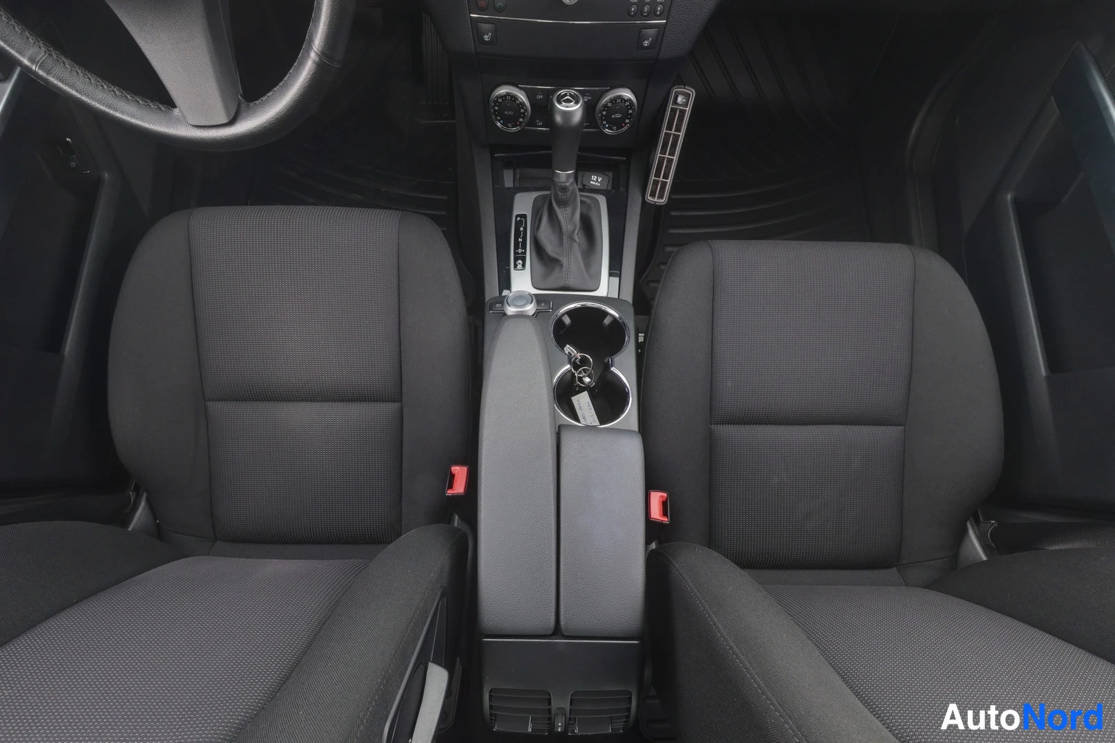 Etuistuimet: Front seats and center console of a car interior with black fabric seats, automatic gear shift, cup holders, and climate control dials on the dashboard.