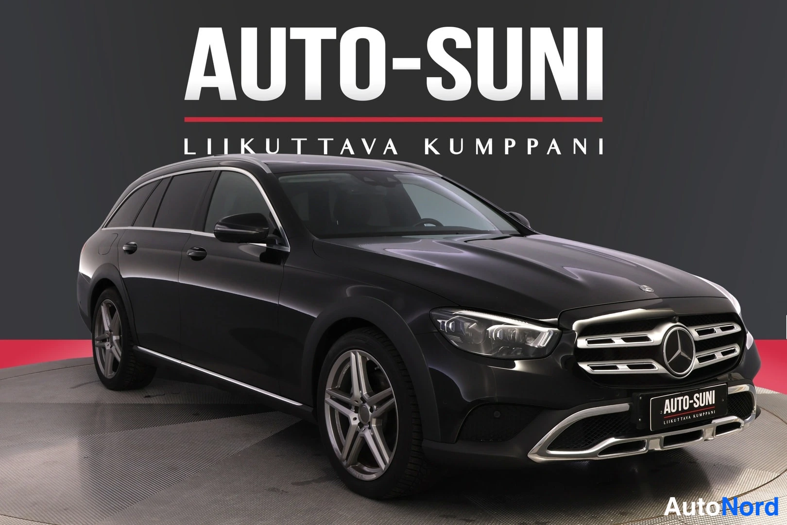 Etuviisto, nokka oikealle: Musta Mercedes-Benz E -farmariauto etuviistosta kuvattuna studiotaustassa, AUTO-SUNI-kyltti taustalla.