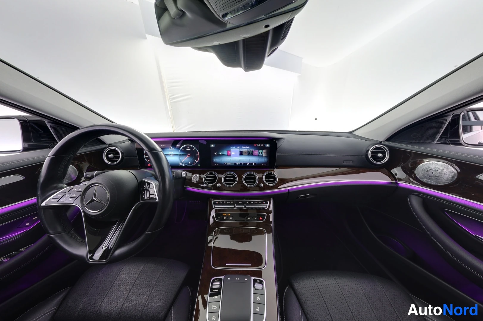 Etuviisto: Ohjaamo etupäältä: Mercedes-Benzin ohjauspyörä, kaksi näyttöä kojelaudassa, pyöreät ilmastointisuuttimet ja violetti tunnelmavalaistus.