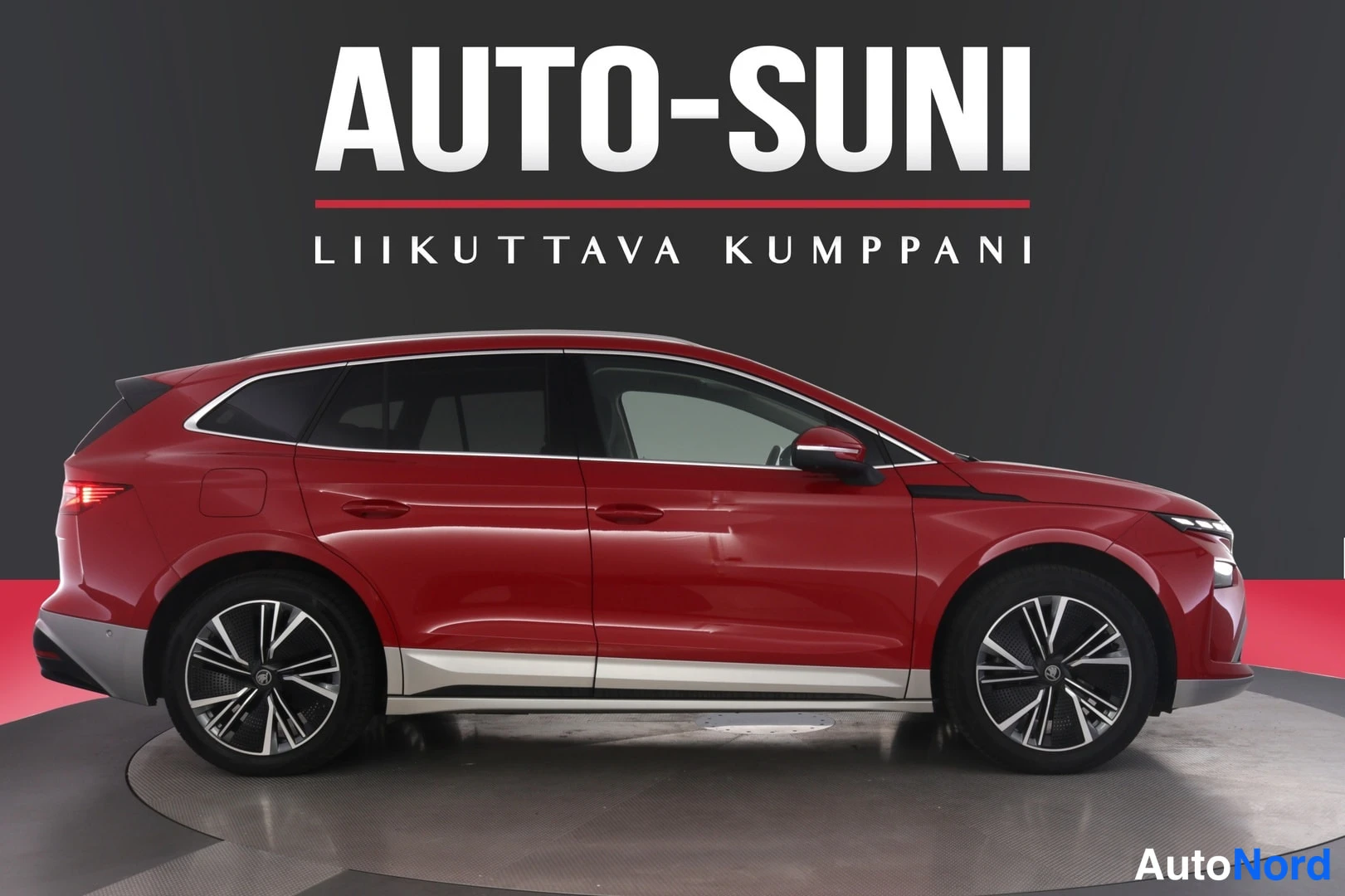 Etuviisto, nokka oikealle: Punainen Skoda Enyaq -farmarimallinen sähköauto sivulta studiotapahtumassa, keula hieman oikealle kääntyneenä, hopeiset alareunan koristeet ja tummat kev