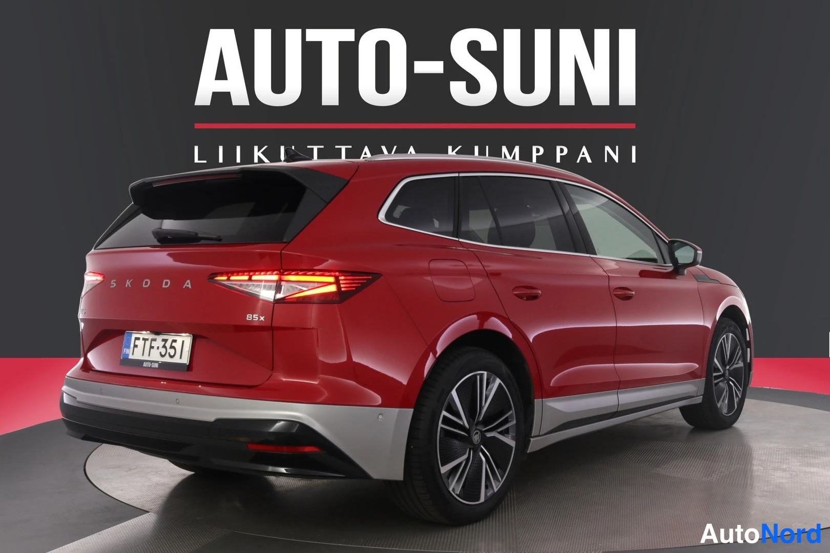 Takaviisto: Punaisen Skoda Enyaqn takaosa kolmannesnäkymästä vasemmalle taaksepäin, SKODA-teksti ja 85x-merkintä takaluukussa, rekisterikilpi, LED-takavalot päällä.