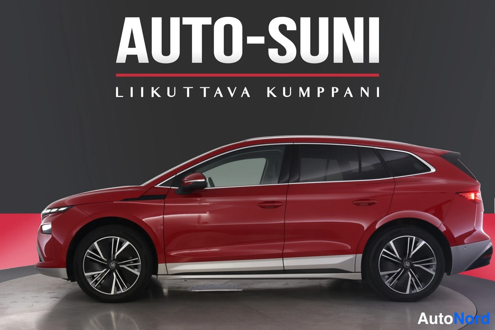 Etuviisto, nokka vasemmalle: Punainen Skoda Enyaq -sähköauto sivulta studiotapahtumassa, keula hieman vasemmalle kääntyneenä, hopeiset alareunan koristeet ja tummat kevytmetallivan